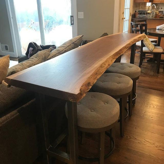 A live edge wood bar top rests on round stools in a cozy home interior, blending rustic charm with modern comfort. Natural light highlights the piece's unique contours and wood grain.